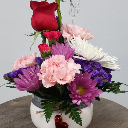 Flower ShopValentine's Day flower arrangement in a white vase with a blue-grey rim and a red heart charm. The bouquet features blooms in shades of light pink, hot pink, red roses, purple, and white, accented with lush greenery for a romantic and elegant look.